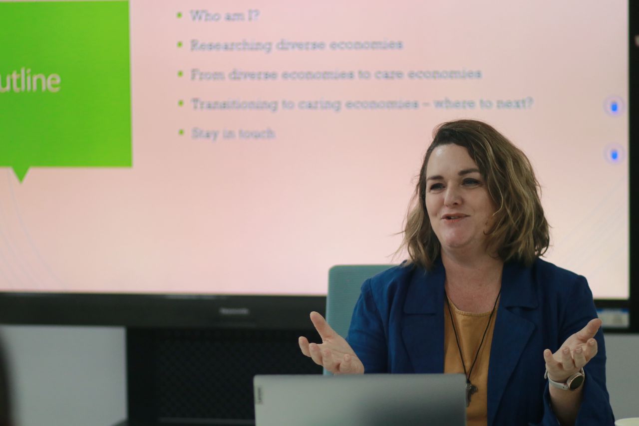 Kelly sits in front of a powerpoint giving a talk